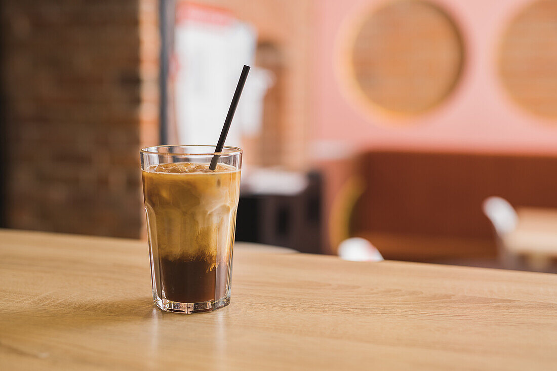 Hohes Glas mit schwarzem Eiskaffee mit sich langsam auflösender Frischmilch und mit Plastikstrohhalm auf dem Tisch in einem hellen Cafe