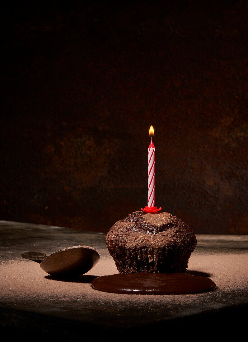 Sweet birthday muffin with powdered sugar and extinguished candle served on table with spilled chocolate and spoon on dark background