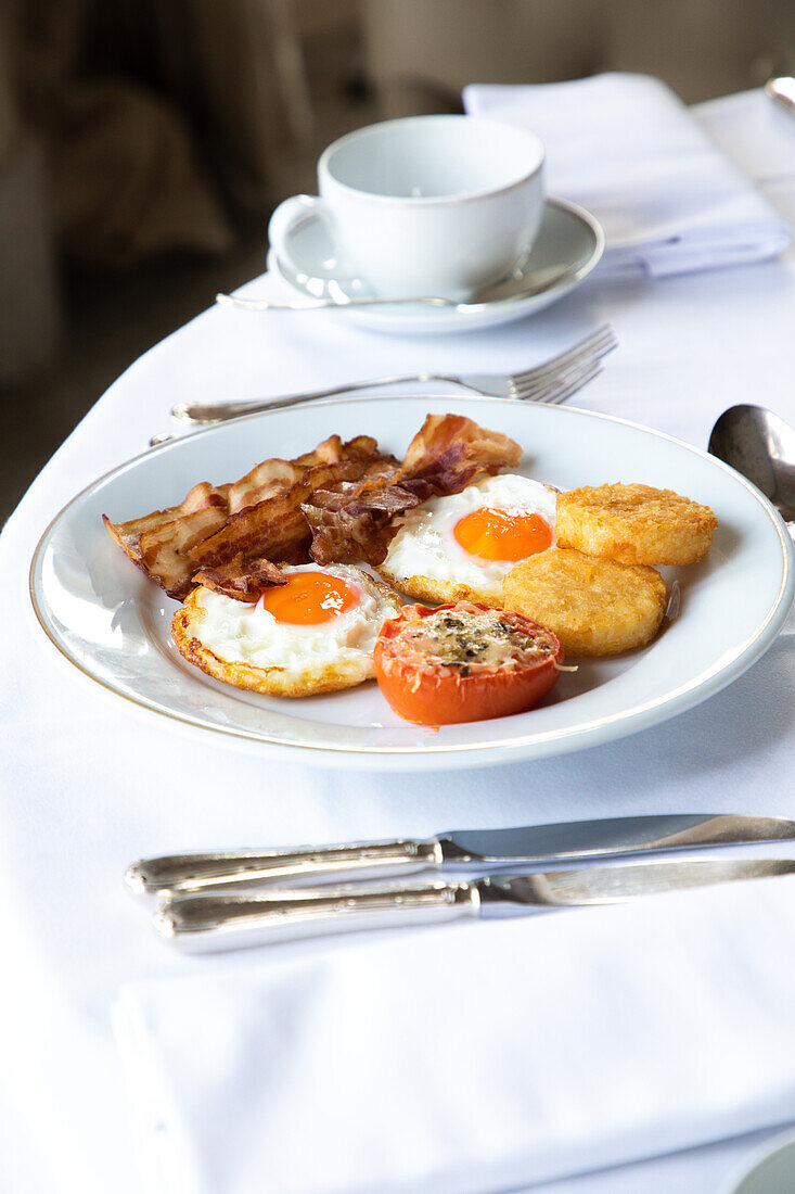 High angle of appetizing fried eggs with bacon slices served on plate with stuffed tomato and crispy mozzarella and placed on table with cutler and cup of coffee during breakfast
