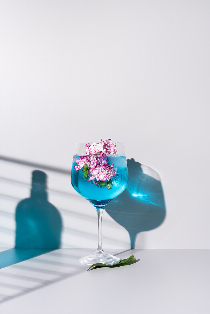 Glasflasche Schatten der blauen Flüssigkeit in der Nähe Glas mit erfrischenden Cocktail mit Eis und Blumen auf dem Tisch vor weißem Hintergrund platziert