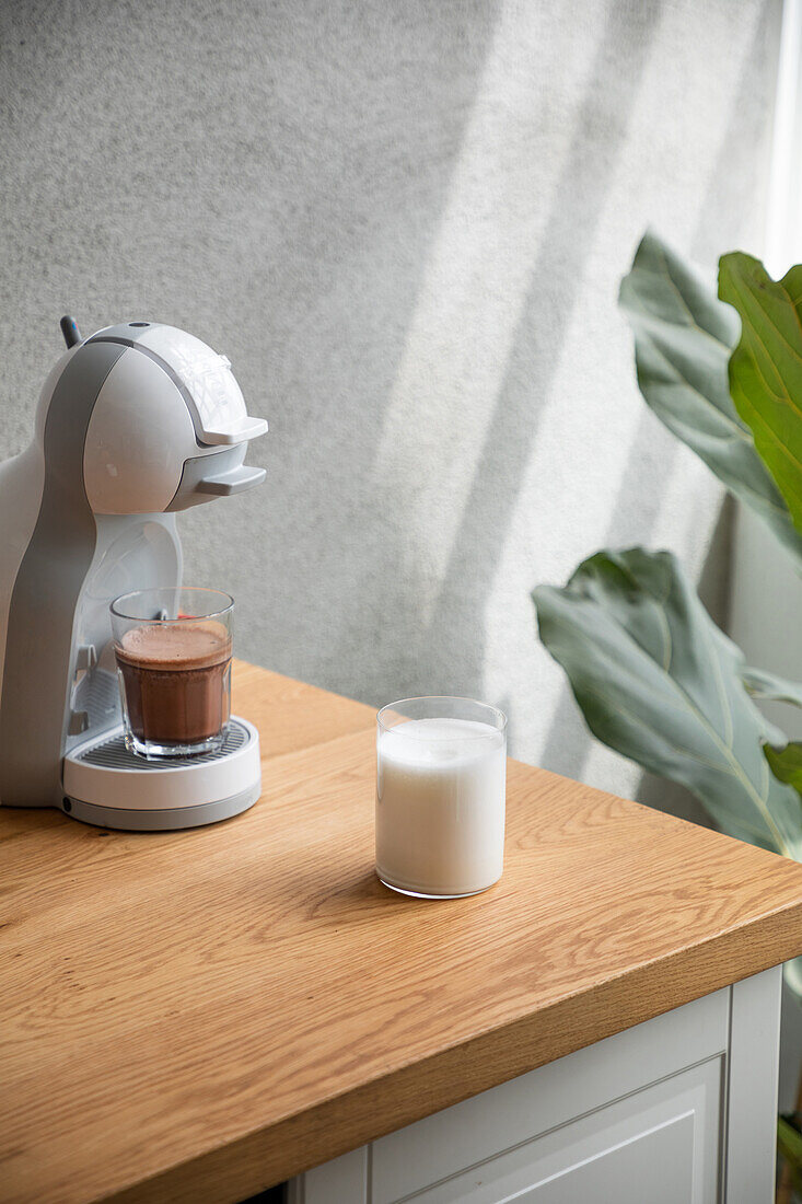 Ein Glas frisch gebrühter Kaffee in einer modernen Kaffeemaschine auf einer Holztheke mit Milch in einer hellen Küche neben einer Grünpflanze