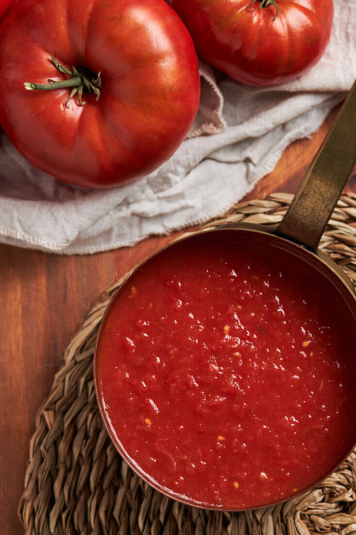 Draufsicht auf eine Pfanne mit köstlicher hausgemachter Tomatensauce, die auf einer gewebten Matte auf einem Holztisch in der Küche steht