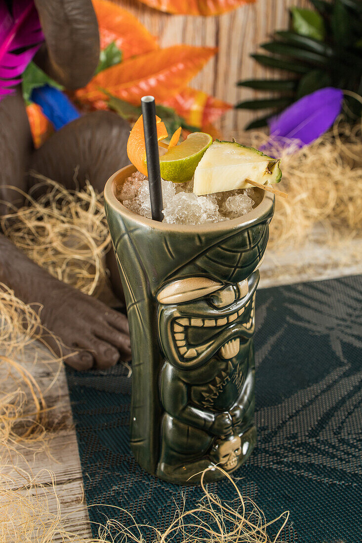 Von oben auf einen großen skulpturalen Tiki-Becher, der mit Alkohol gefüllt und mit Stroh und Früchten verziert ist und auf einem grünen Teppich im trockenen Gras steht