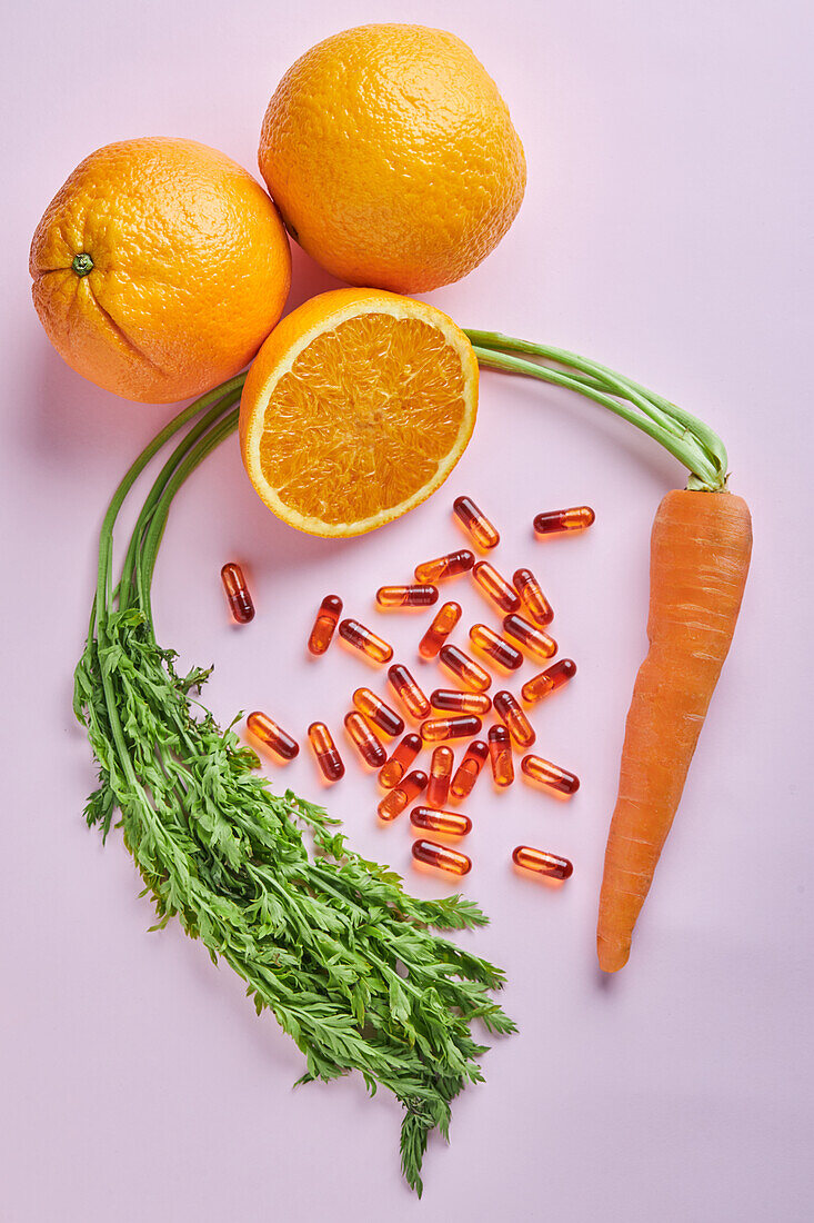 From above composition of scattered vitamin pills arranged on pink table near ripe carrots and juicy oranges