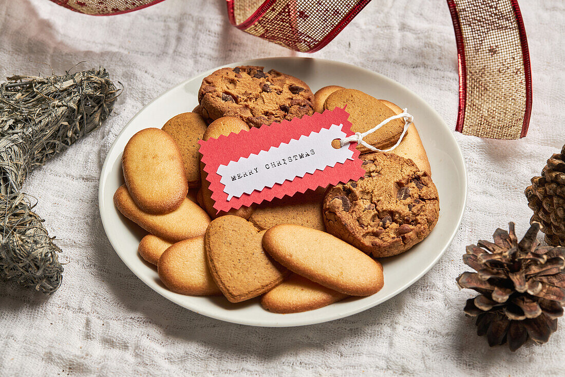 Teller mit verschiedenen süßen Keksen und Geschenkanhänger auf einem Tisch mit Weihnachtsdekoration von oben