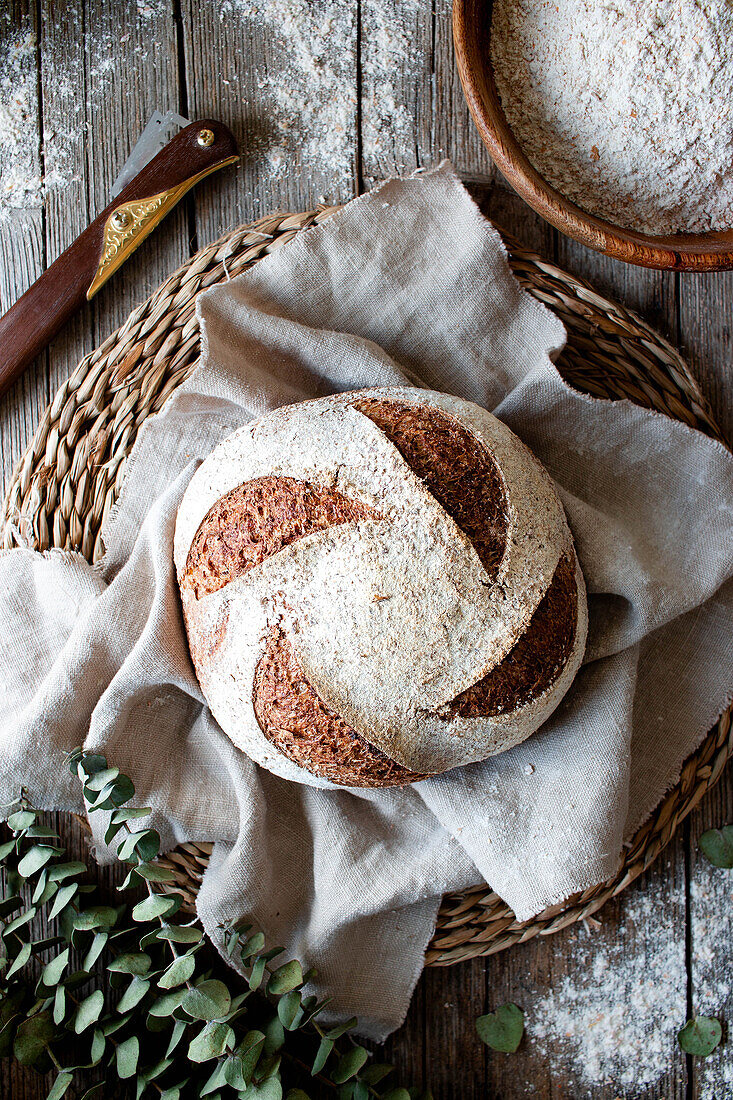 Von oben hausgemachtes frisches Sauerteigbrot Dinkelbrot auf Weidenständer mit Tuch auf Holztisch mit Mehl bestreut