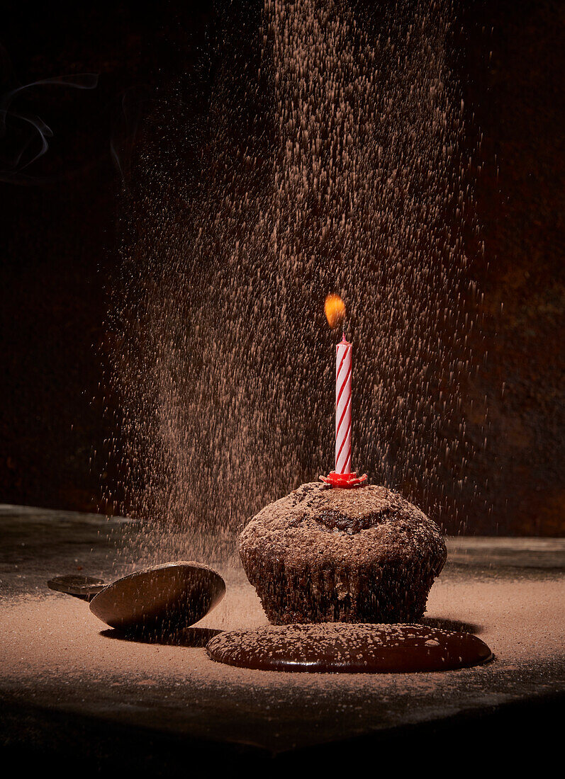 Powdered sugar sprinkling on tasty sweet birthday muffin with powdered sugar and extinguished candle served on table with spilled chocolate and spoon on dark background