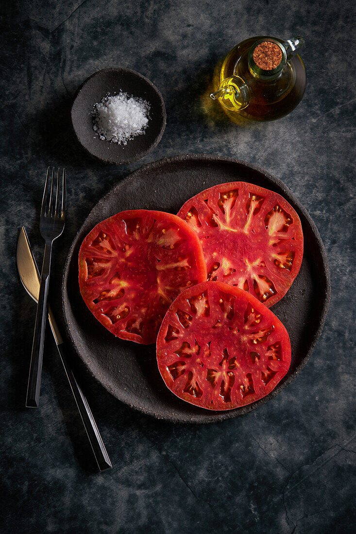 Top view of delicious sliced tomatoes in cast iron plate near sea salt and jug of olive oil on concrete table