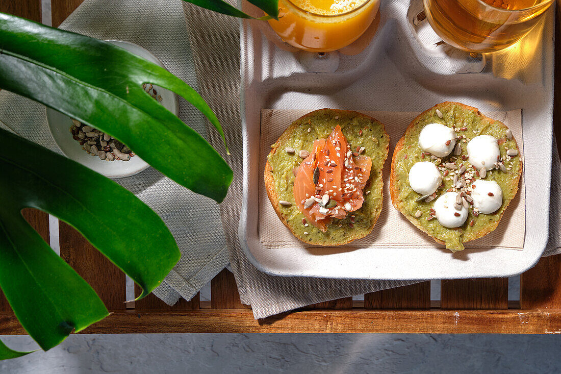 From above of delicious avocado toasts with salmon and burrata cheese served on table with glasses of fresh juice and herbal tea placed near exotic Monstera deliciosa plant