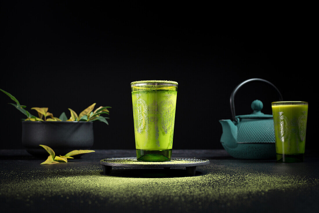 Still life composition with traditional oriental matcha tea served in glass cup with metal ornamental decor on table with ceramic bowls and fresh green leaves against black background
