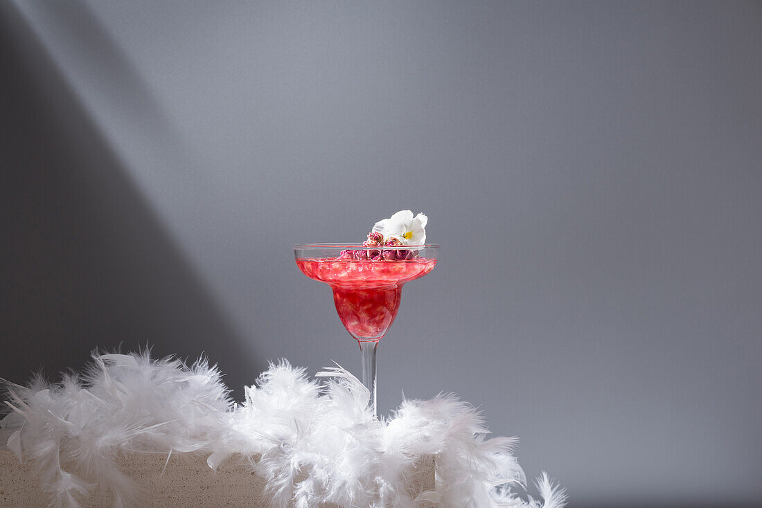 Crystal glass of pomegranate margarita cocktail served with flower blooms near near feathers on concrete blocks in studio