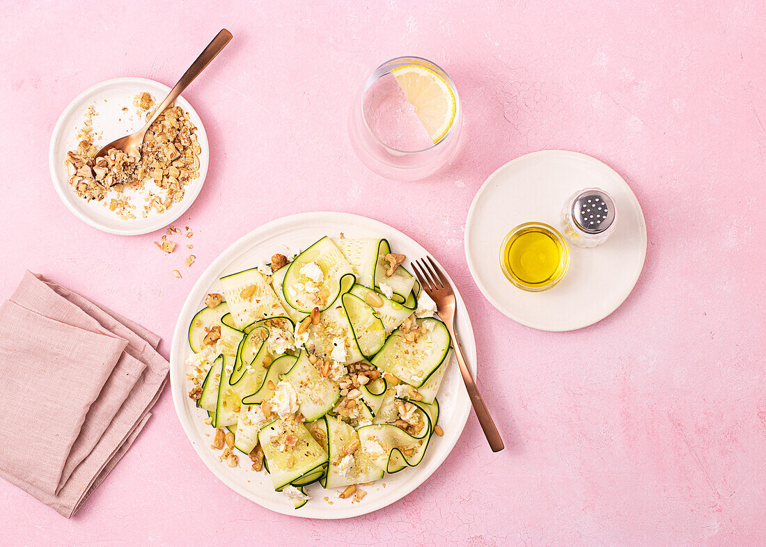 Frischer Salat aus dünn geschnittenen reifen Zucchini, Fetakäse und Pinienkernen in einem weißen Teller auf einem rosa Hintergrund