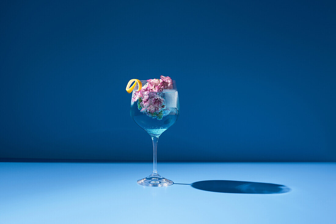 Transparent glass of fresh cocktail with mint leaves and flowers placed on surface against blue background