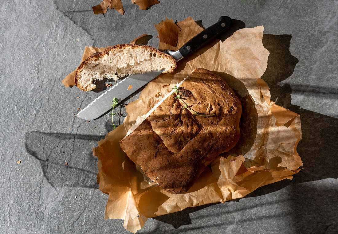 Draufsicht auf ein köstliches, frisch gebackenes Sauerteigbrot mit aufgeschnittenem Stück und Messer auf Backpapier auf grauem Hintergrund