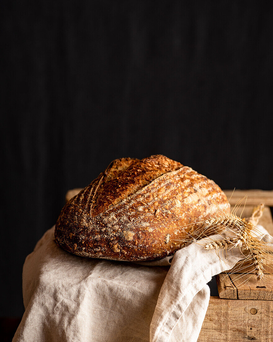 Delicious whole artisan bread with crust on crumpled fabric against wheat spikes on wooden surface