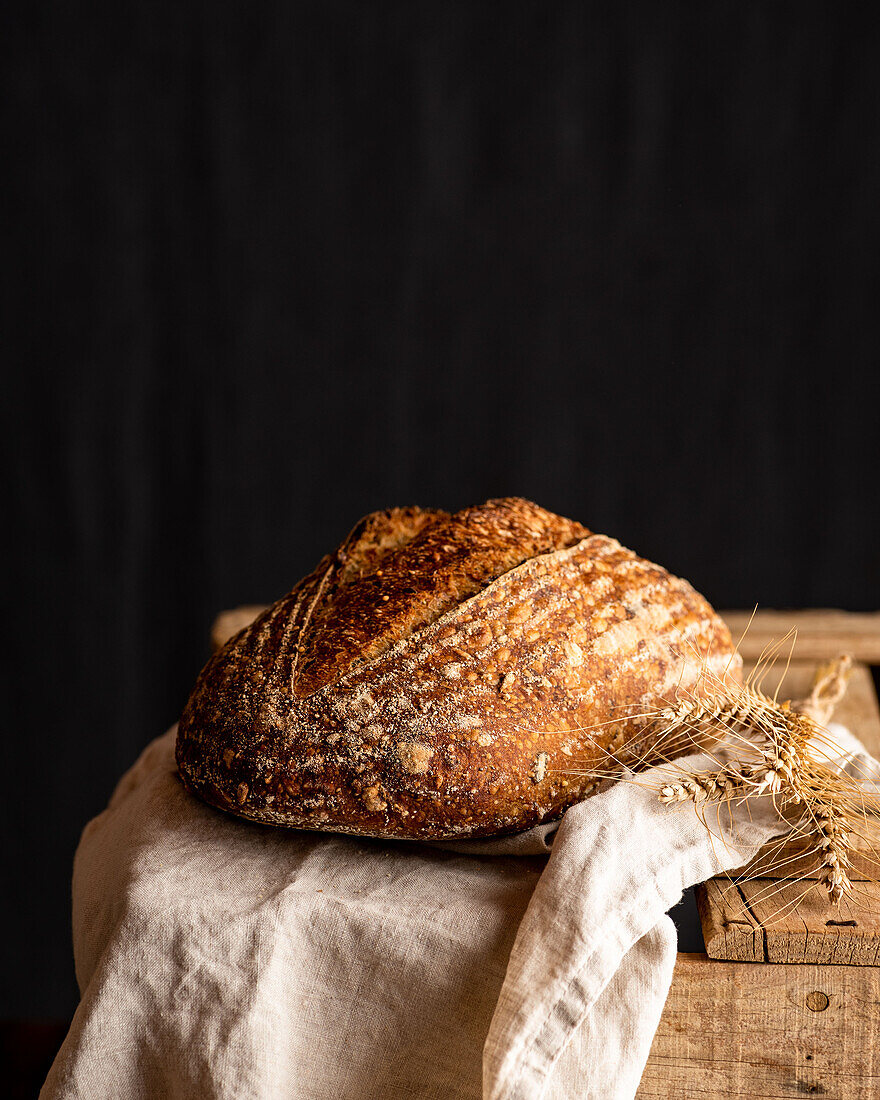 Leckeres handwerkliches Vollkornbrot mit Kruste auf zerknittertem Stoff gegen Weizenspitzen auf Holzoberfläche