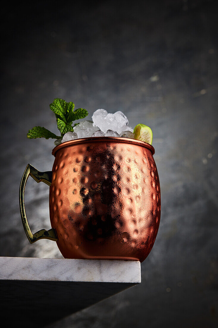 Moscow-Mule-Cocktail, serviert in einem Becher mit Eis und einer Limettenscheibe in einer Tischecke vor grauem Hintergrund