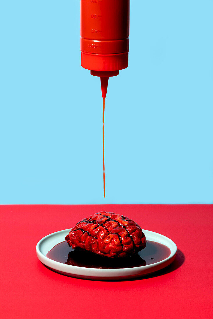 Falsches Blut tropft aus roter Ketchupflasche auf ein Gehirn auf einem Teller mit zweifarbigem Hintergrund
