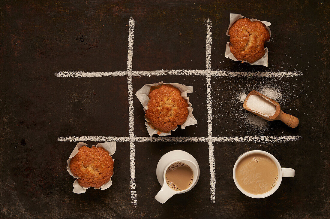 Draufsicht auf ein essbares Tic-Tac-Toe-Spiel mit gebackenen Muffins und einem Löffel mit Zucker und einer Tasse Milchkaffee, die ein Viereck darstellen, auf schwarzem Hintergrund