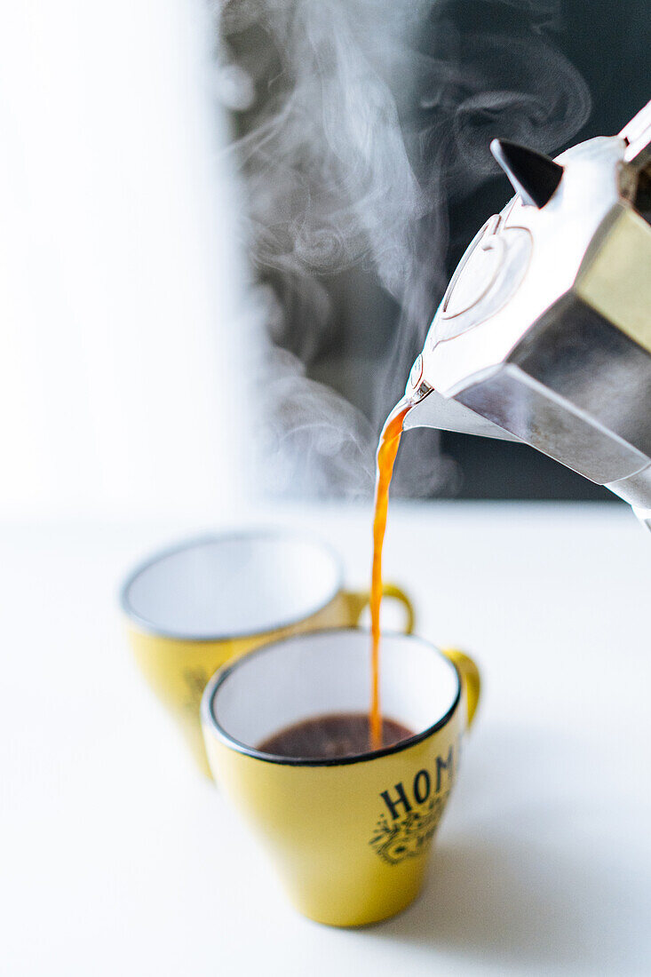 Freshly brewed steaming hot coffee pouring from metal moka pot into yellow cup on table in light kitchen on blurred background
