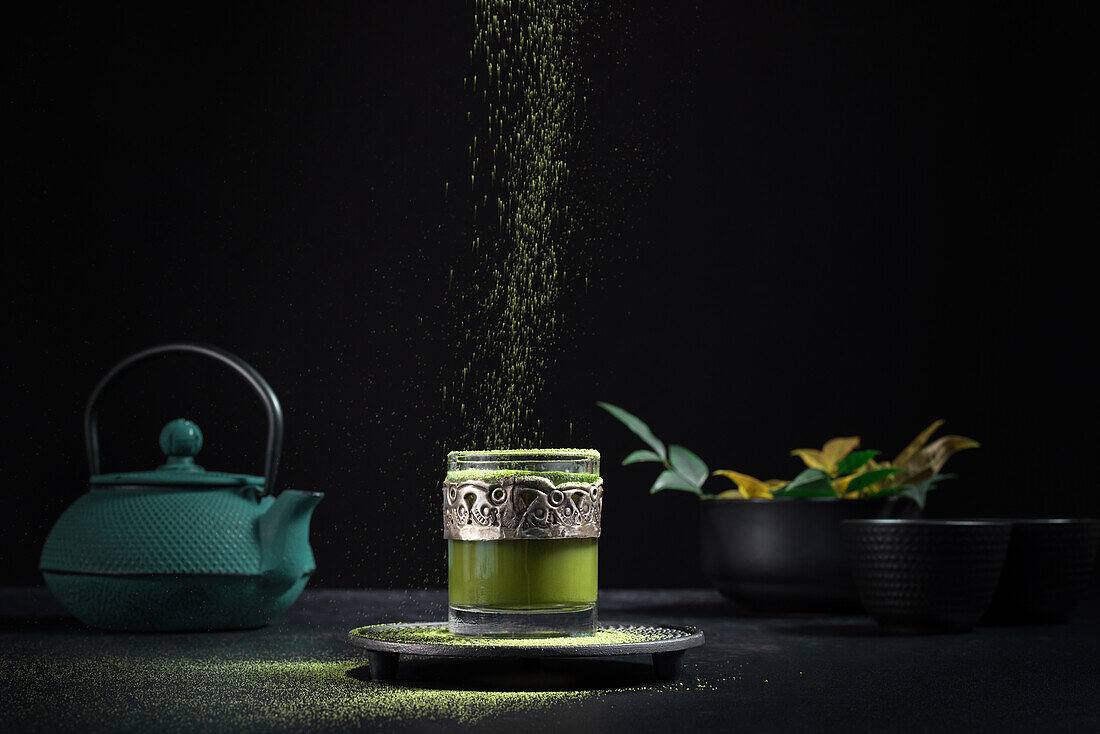 Still life composition with traditional oriental matcha tea served in glass cup with metal ornamental decor on table with ceramic bowls and fresh green leaves against black background