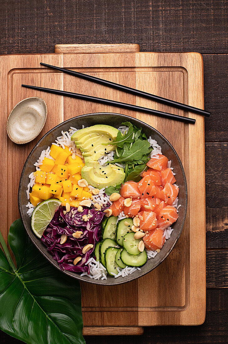 Top view of appetizing colorful poke bowl with fresh salmon and rice served with ripe avocado slices, cabbage and mango placed on wooden tray