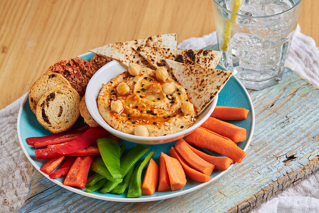 Leckerer Hummus mit Tortilla-Chips auf einem Teller mit Croutons und verschiedenen Gemüsesorten auf einem rustikalen Schneidebrett neben einem Glas kalter Limonade auf einem Holztisch mit Serviette von oben