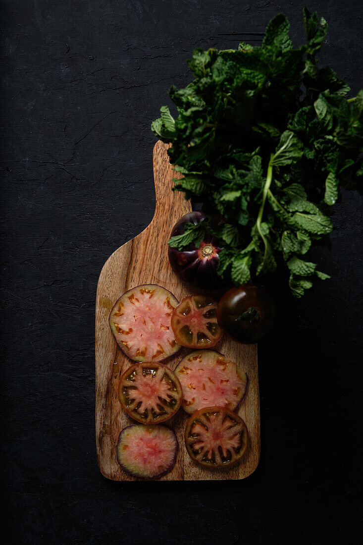 Draufsicht auf frische, reife, in Scheiben geschnittene schwarze Tomaten und grüne Minzstängel auf einem hölzernen Schneidebrett auf schwarzem Hintergrund