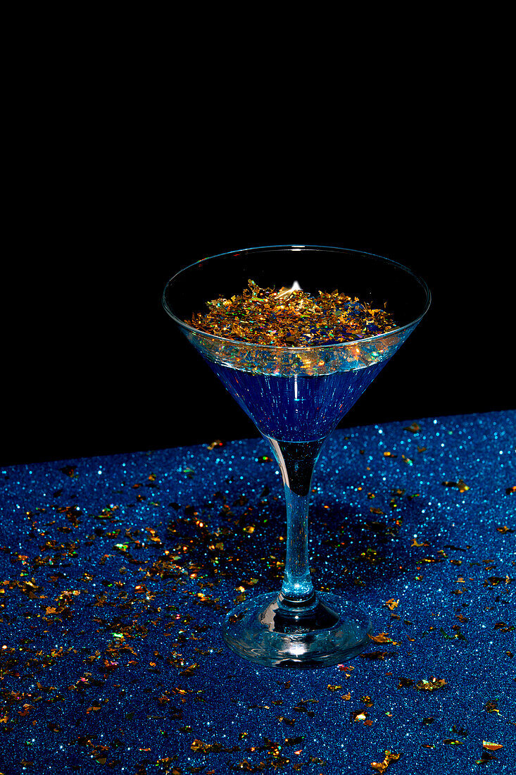 High angle of transparent glass of martini with gold flakes placed on blue counter against black background