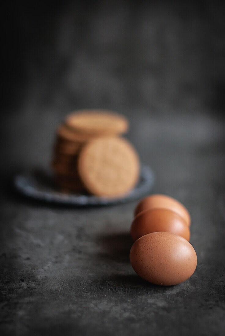 Closeup of raw eggs placed on gray surface near tray with pile of sweet delicious digestive biscuits