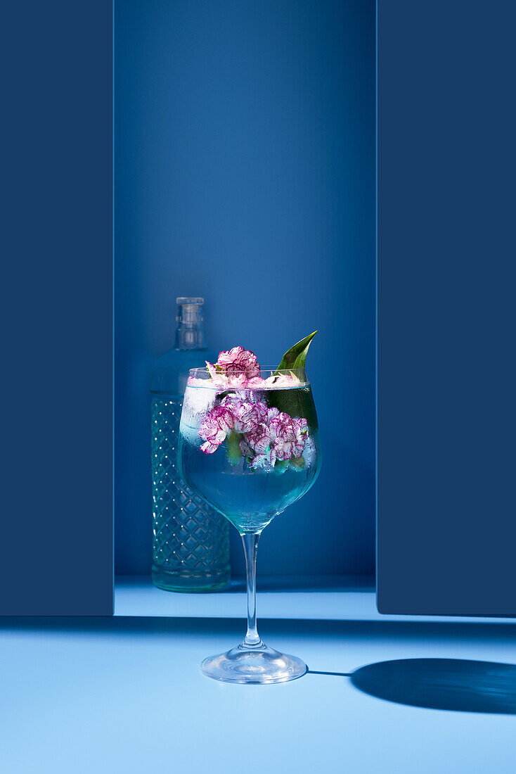 Transparentes Glas eines frischen Cocktails mit Minzblättern und Blumen auf der Oberfläche neben der Flasche vor blauem Hintergrund