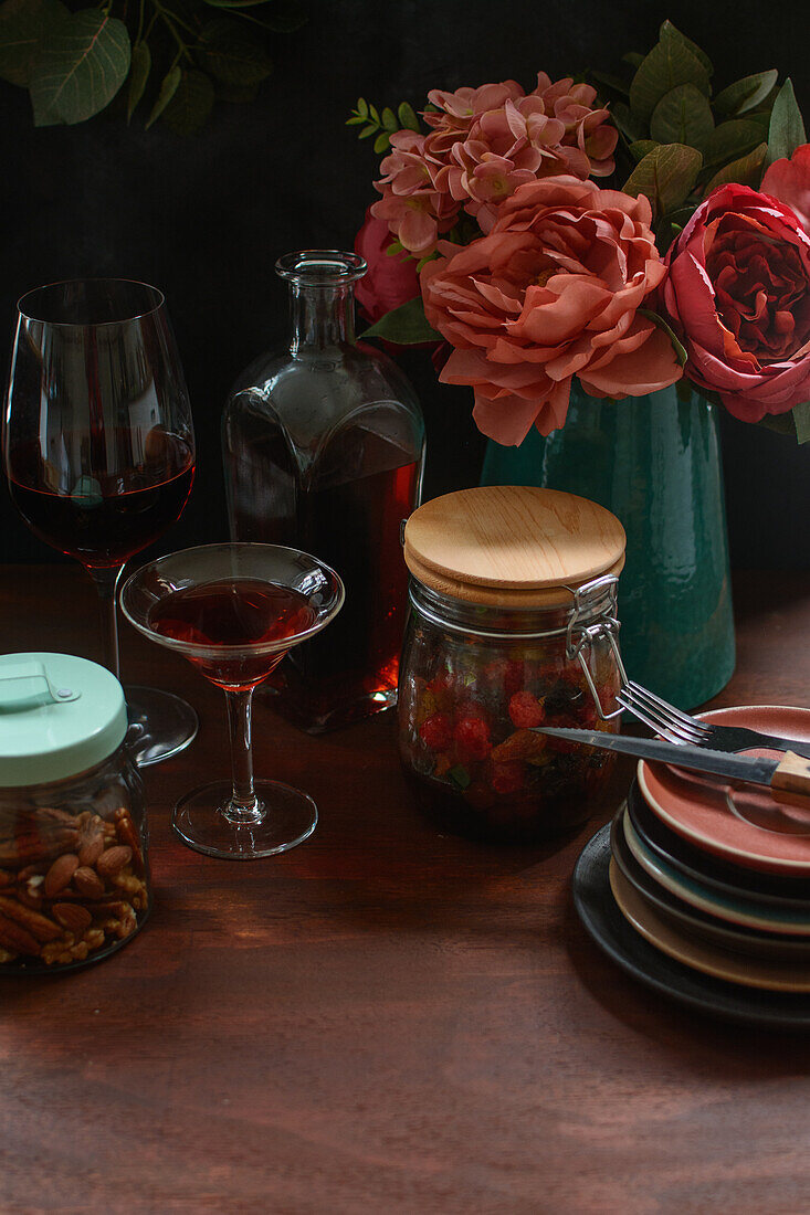 Glass jars with nut and berries on wooden table with tableware and alcohol drink near vase with blooming flowers in kitchen