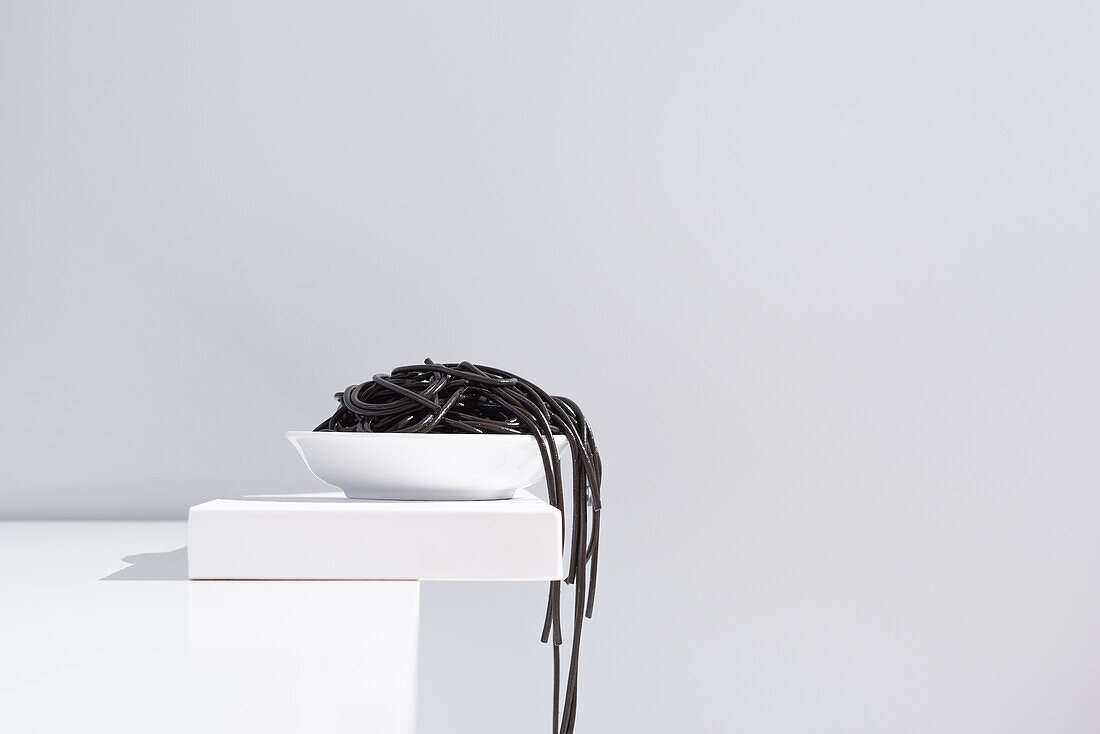 Minimalist studio with black squid ink spaghetti falling out from full ceramic bowl on white table