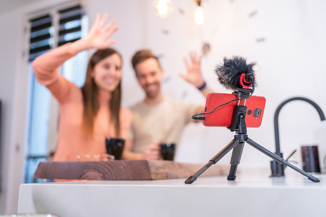 Positives Bloggerpaar, das mit der Hand in ein Handy mit Mikrofon winkt, während es zu Hause am Küchentisch mit einer Tasse Kaffee sitzt