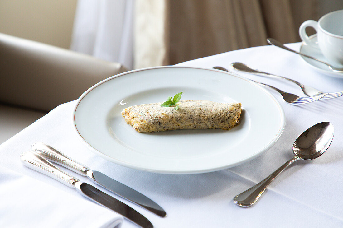 From above of delicious sweet rolled pancake with chocolate and mint leaf placed on white plate served on table white coffee cup and cutlery during breakfast in restaurant