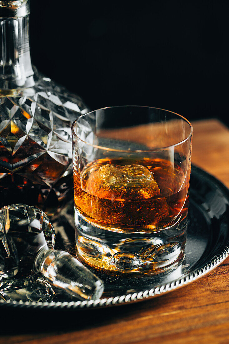 Glass of cold whiskey with ice placed on wooden table near decanter on black background