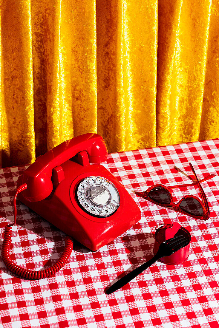 Blick von oben auf ein rotes Retro-Telefon und eine altmodische weibliche Sonnenbrille in der Nähe einer Dose mit einer Gabel auf einem karierten Tischtuch