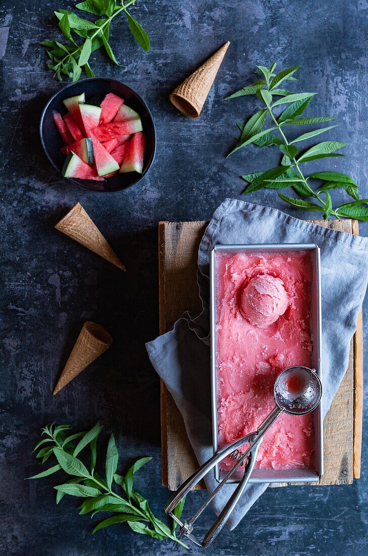Top view of container with sweet watermelon sorbet and scoop placed on napkin and board near mint twigs and cornets on gray table