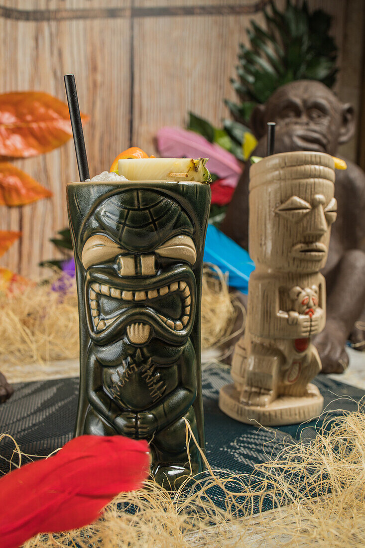 Großer skulpturaler Tiki-Becher, gefüllt mit Alkohol, verziert mit Stroh und Früchten, auf einem grünen Teppich vor trockenem Gras