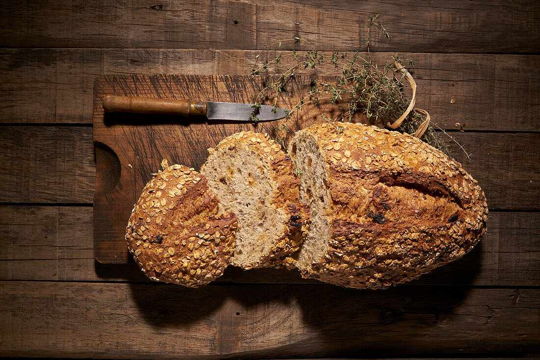 Top view of appetizing freshly baked homemade natural bread loaf with crispy crust and oat flakes on wooden table with knife and aromatic herbs