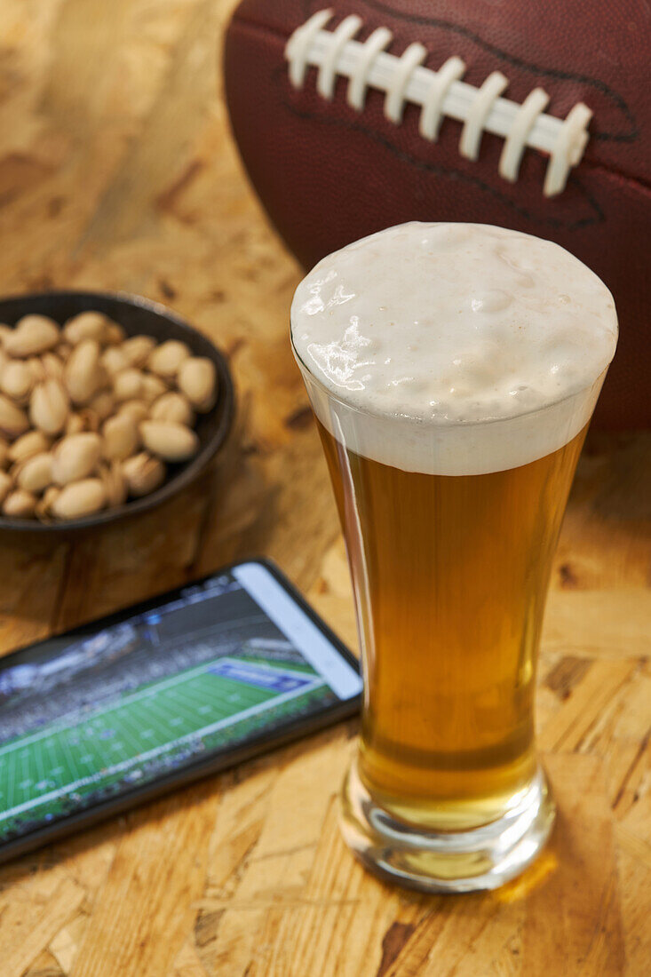High angle of glass of fresh cold beer placed on wooden table near plate with pistachios and ball before watching match on cellphone