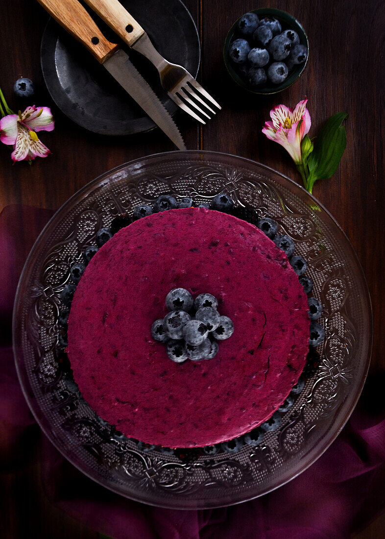 Köstliche Blaubeer-Mousse-Torte mit violetter Sahne, dekoriert mit frischen Beeren, serviert auf einem Glasständer auf einem dunklen Tisch