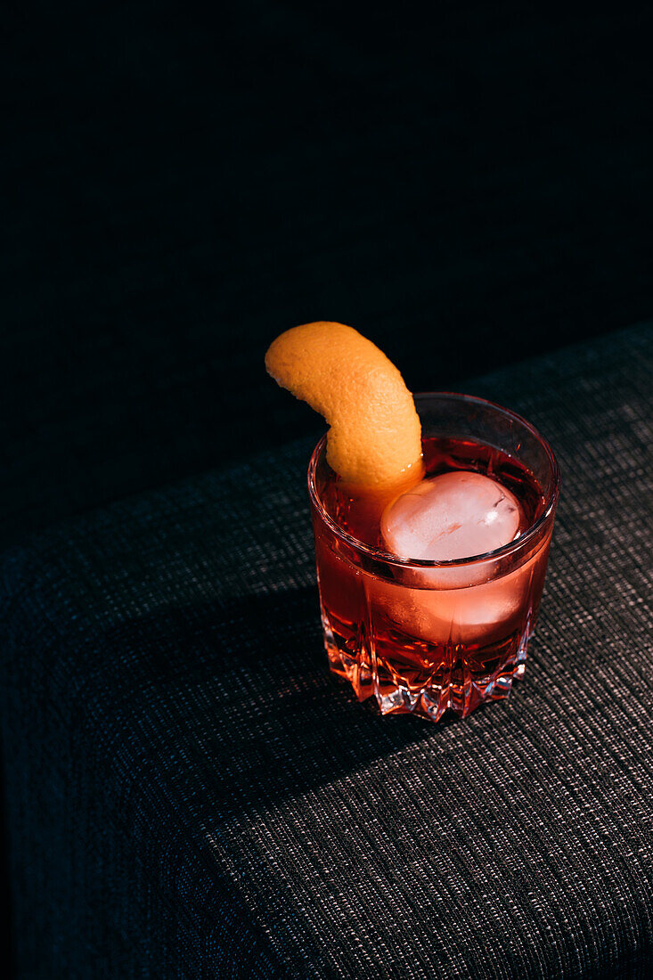 Glass of refreshing Negroni cocktail with bitter flavor and ice garnished with orange peel and served on couch arm in dark room