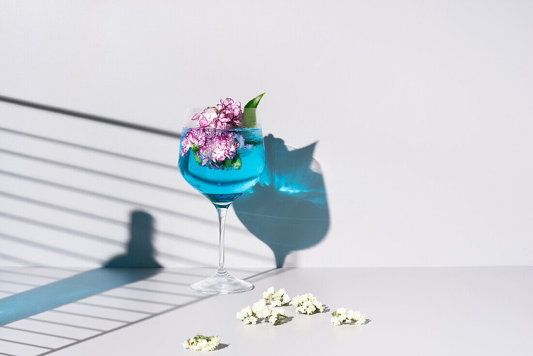 Glass bottle shadow of blue colored liquid placed near glass with refreshing cocktail with ice and flowers on table against white background