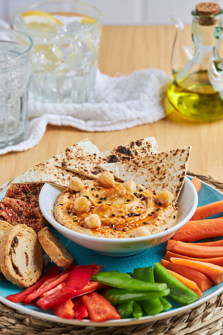 Schale mit Hummus und Tortilla-Chips, umgeben von verschiedenen Gemüsesorten und Croutons, serviert auf einer Weidenmatte neben Olivenöl und Limonade auf einem Holztisch von oben