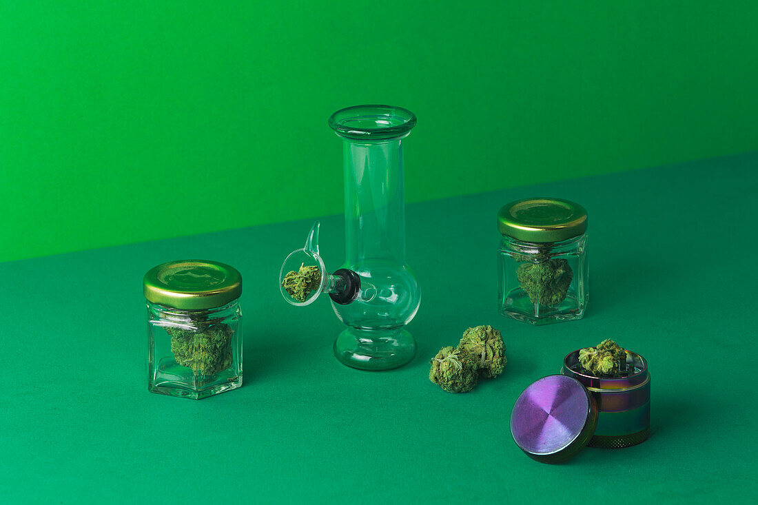Composition of glass round bong placed near dry cannabis plant in bowl on green surface