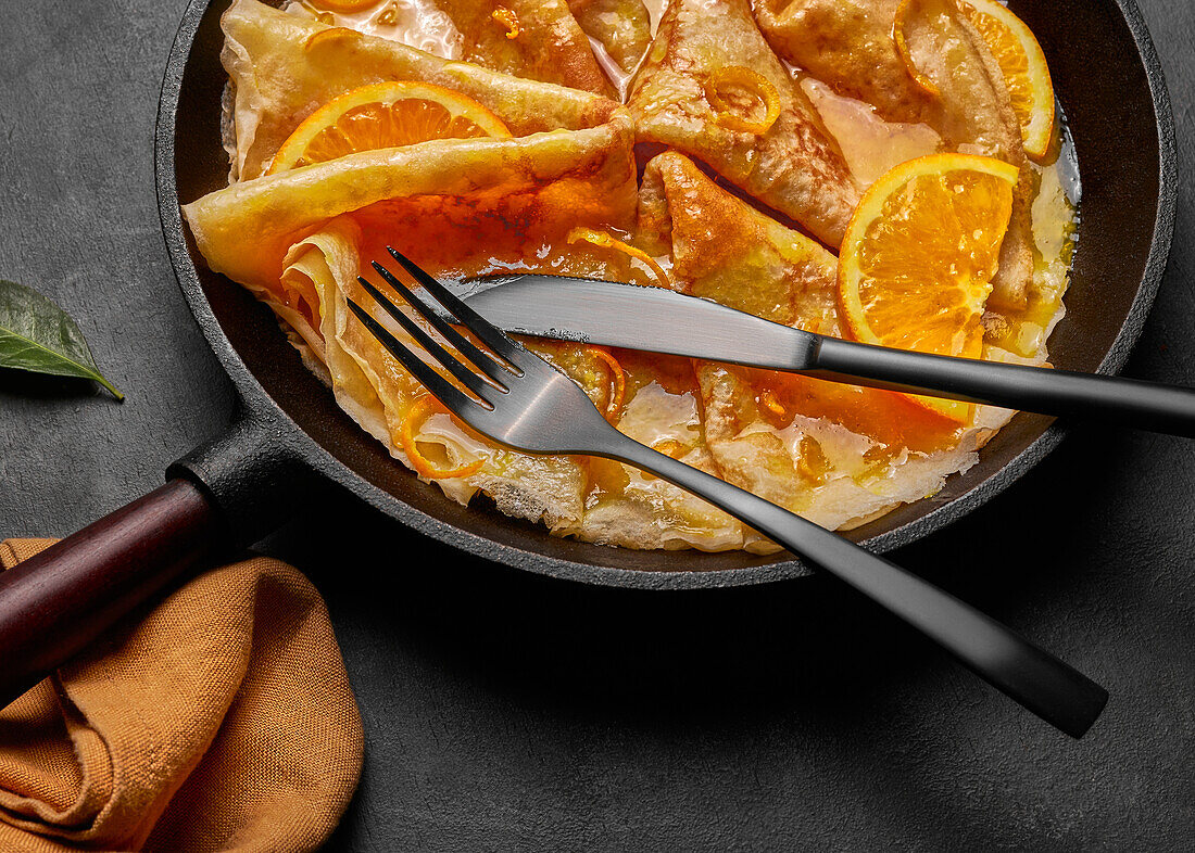 From above homemade crepes suzette with orange liqueur and orange slices served on plate with knife and fork on concrete surface