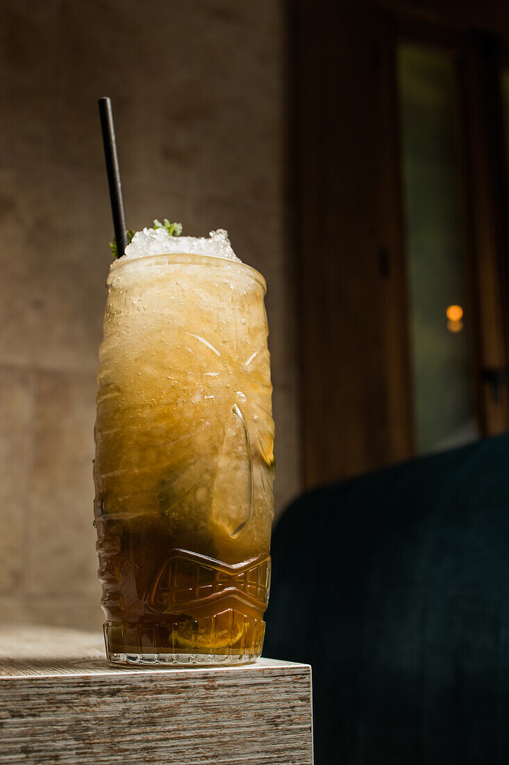 Tiki cup with cold alcohol drink with straw served with ice and decorated with fresh herb placed on blurred background