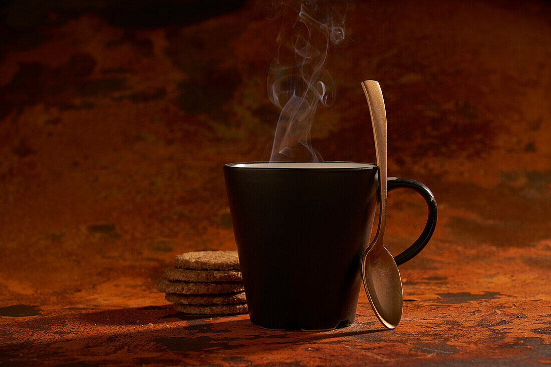 Dark ceramic mug of hot aromatic drink and spoon place on surface near pile of fresh baked oatmeal cookies