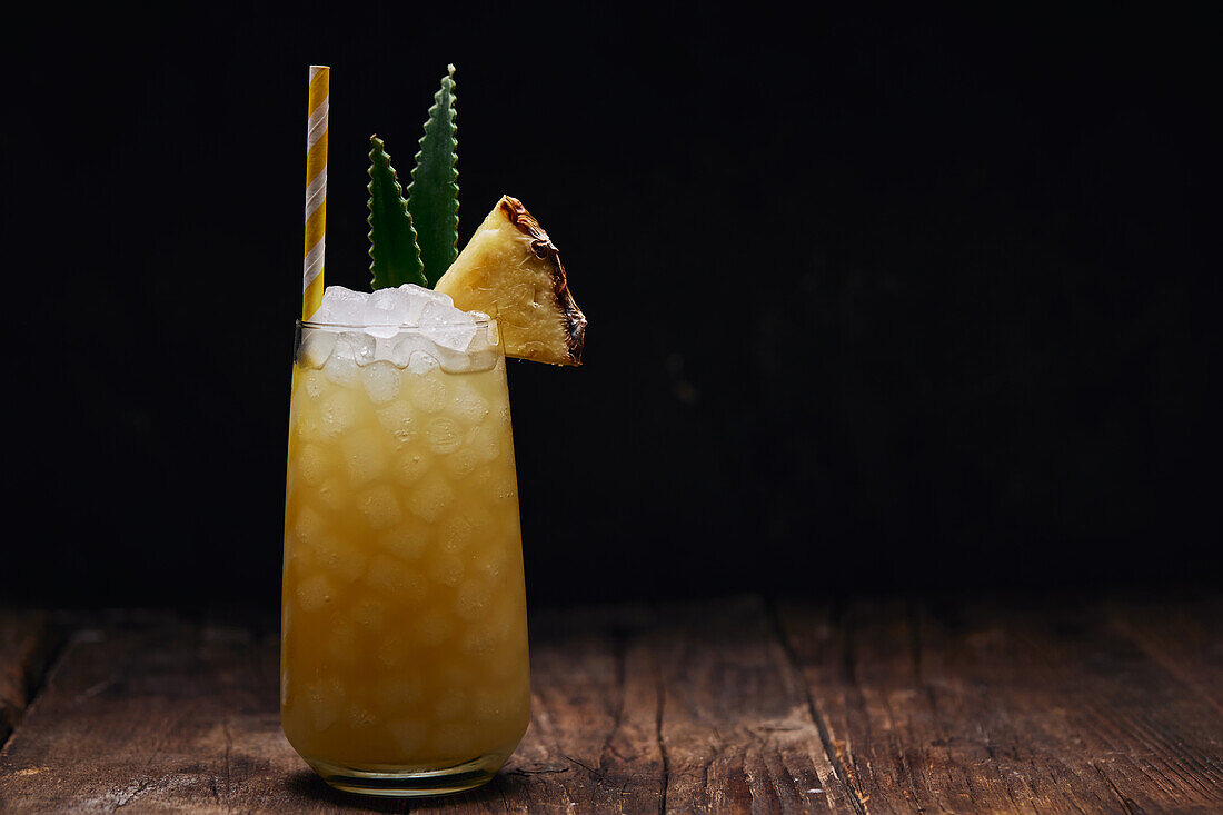 Wooden table with glass of yellow cocktail with ice cubes and refreshing cocktail garnished with spiky leaves and striped straw